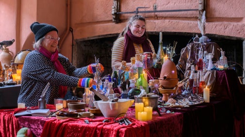 A table covered with coloured potions in glass bottles, fairy lights and earthenware bowls is in the foreground, with two women dressed in winter clothing behind.